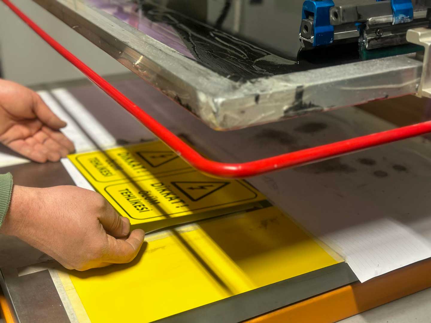 Serigrafi baskı işlemi sırasında bir kişi, mürekkebi serigrafi çerçevesi üzerinden sıyırıcı ile geçirerek baskı yapıyor